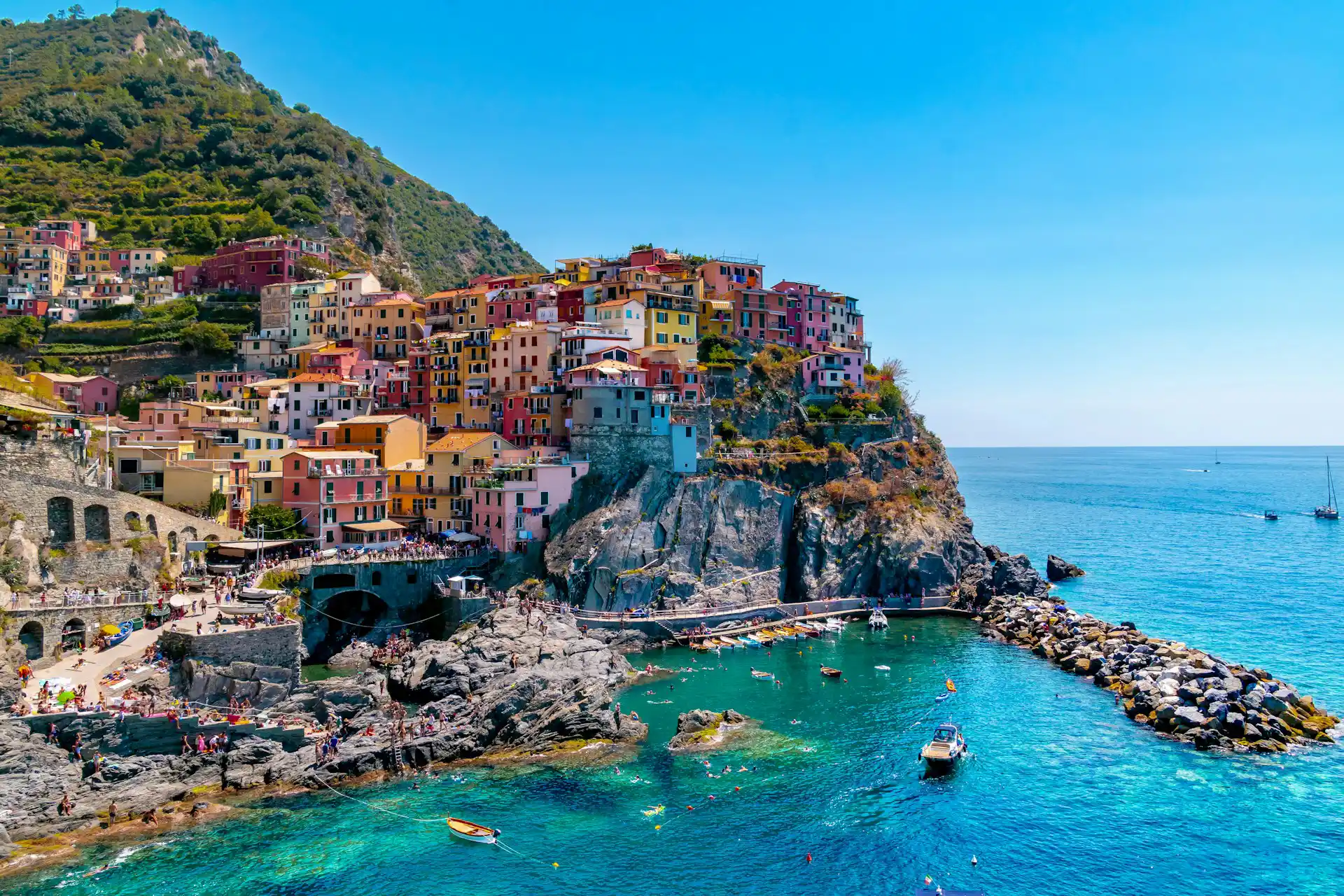 Il paese di Manarola nelle Cinque Terre con case color pastello affacciate su una piccola insenatura dal mare turchese. In primo piano si vedono barche ormeggiate e una scogliera rocciosa, mentre sullo sfondo le colline verdi degradano verso il mare sotto un cielo azzurro limpido.