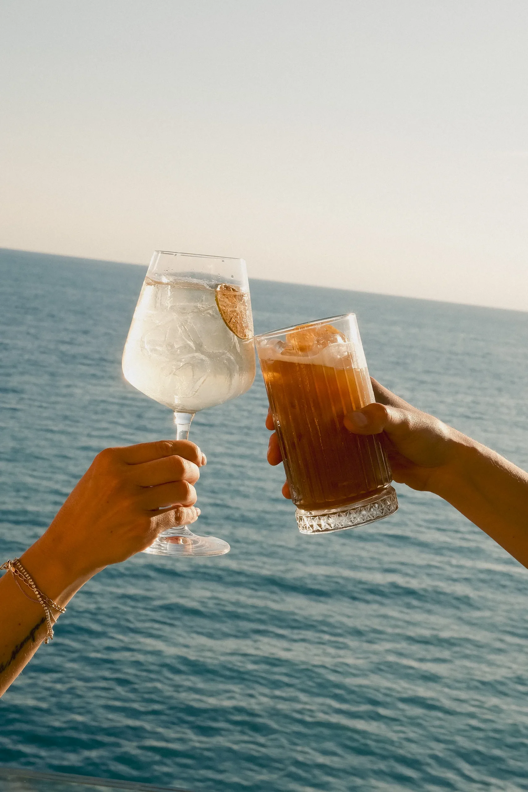Due mani che brindano davanti al mare aperto, sollevando un calice con cocktail chiaro decorato con fetta di agrume e un bicchiere con drink ambrato e ghiaccio. Sullo sfondo si estende l’acqua blu fino all’orizzonte, illuminata dalla luce calda del sole.