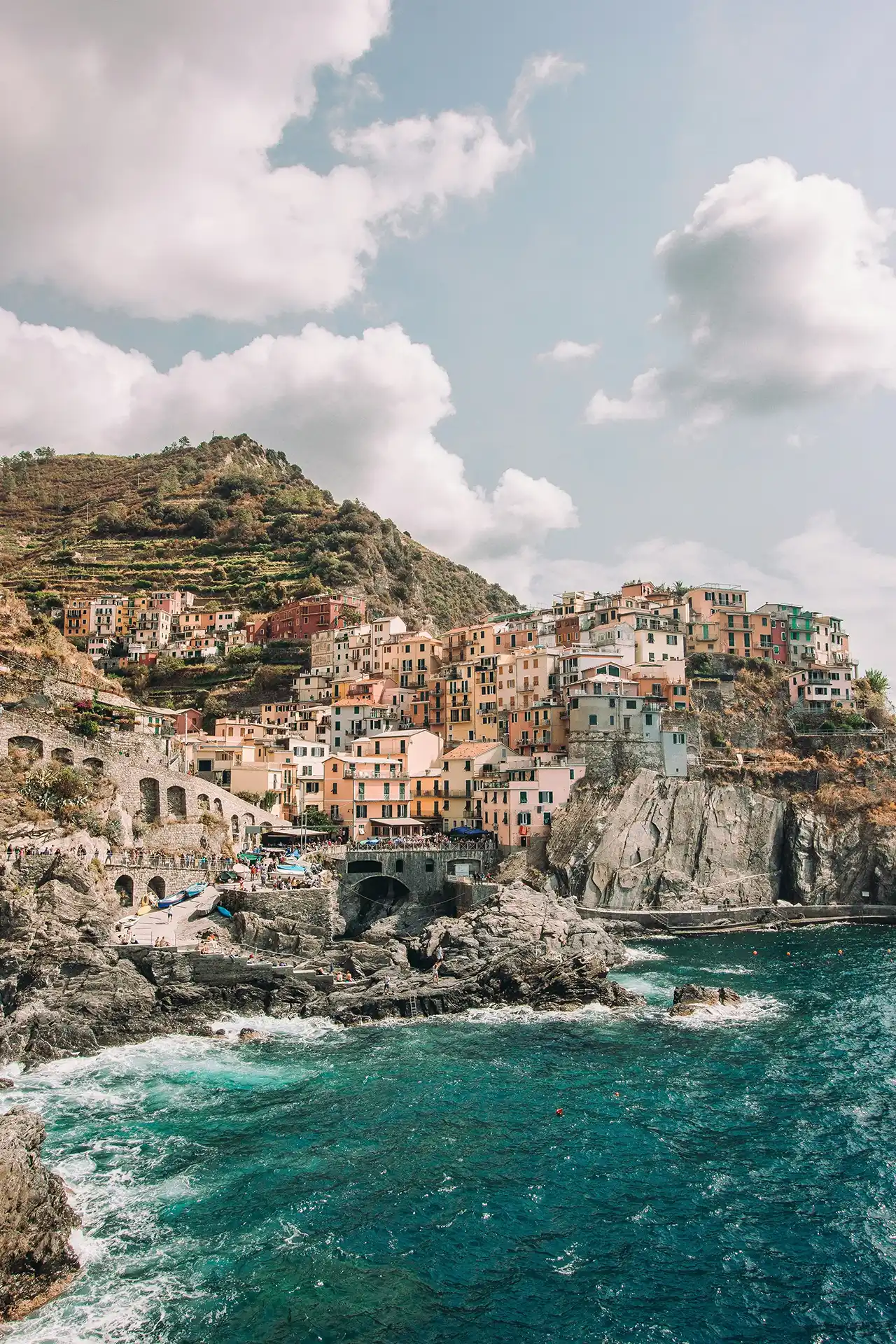 Il borgo di Manarola su una scogliera della Cinque Terre con case color pastello disposte a terrazze sopra il mare. In primo piano l’acqua turchese si infrange sulle rocce. Sullo sfondo, colline verdi e cielo parzialmente nuvoloso creano una luce morbida sulla scena.