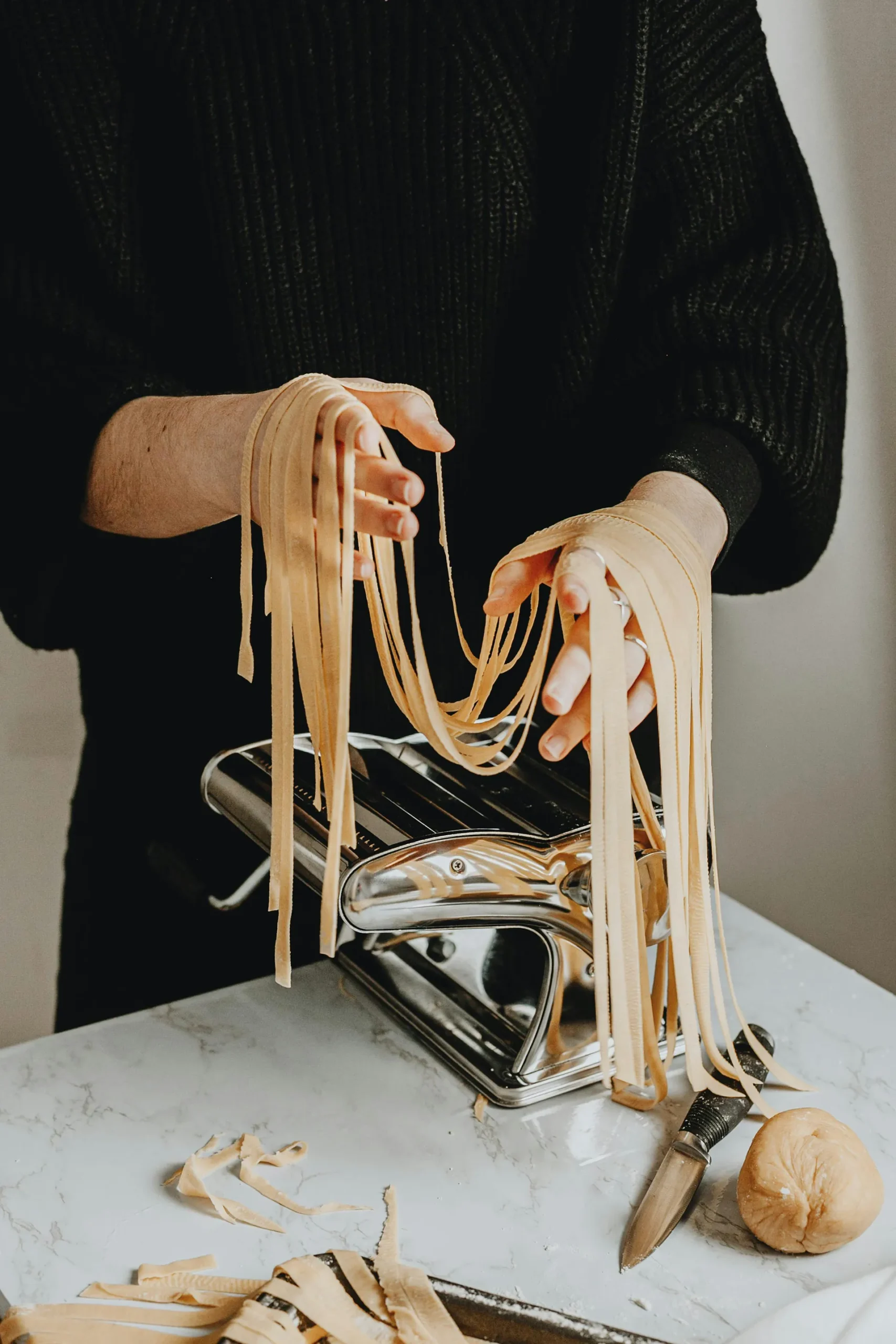 Mani che lavorano la pasta fresca con una macchina manuale, mentre lunghe strisce di pasta all’uovo fuoriescono dai rulli. In primo piano si vedono la sfogliatrice in metallo, un coltello e una piccola porzione di impasto su un piano in marmo chiaro.
