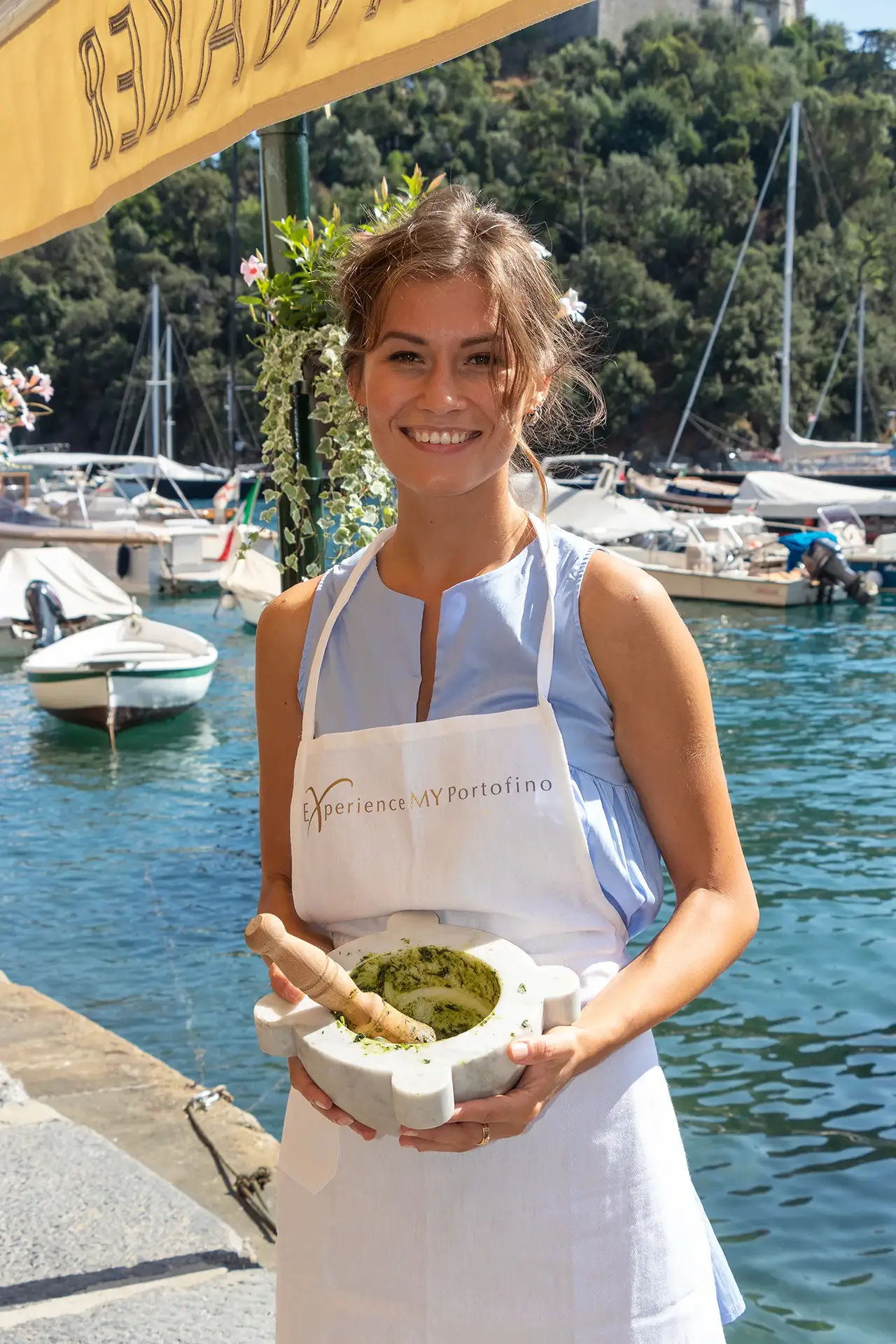 Ragazza in piedi su un molo con vista su un porto turistico, mentre tiene tra le mani un mortaio in marmo con pesto verde e pestello in legno. Indossa un grembiule chiaro con la scritta “Experience MY Portofino”. Sullo sfondo si vedono barche ormeggiate, acqua azzurra e una collina verde.