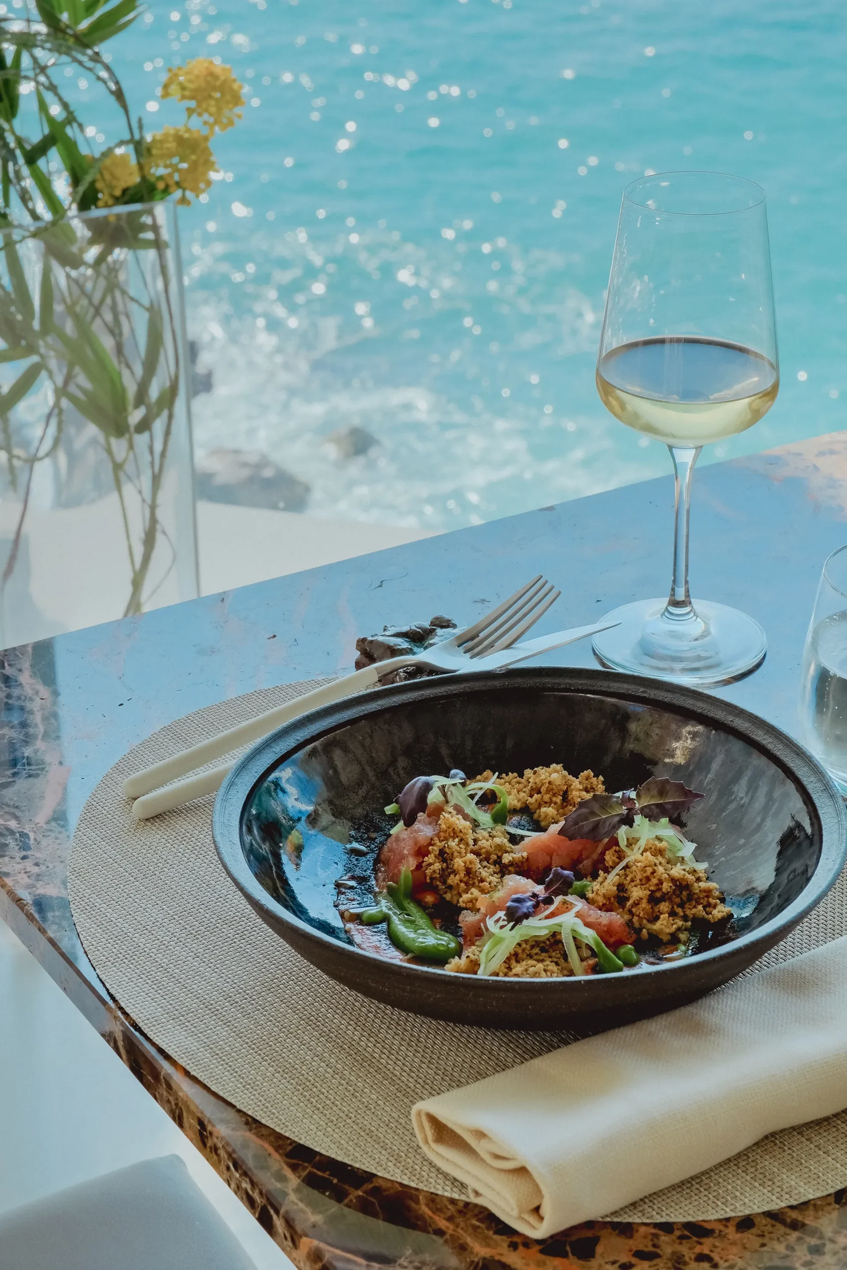 Tavolo del ristorante apparecchiato vicino a una vetrata affacciata sul mare, con un piatto scuro che presenta una pietanza guarnita con verdure e granelle croccanti. Accanto si vedono un calice di vino bianco, posate su tovagliolo piegato e un vaso con fiori gialli. Sullo sfondo, l’acqua turchese del mare riflette la luce naturale creando un’atmosfera luminosa e raffinata.