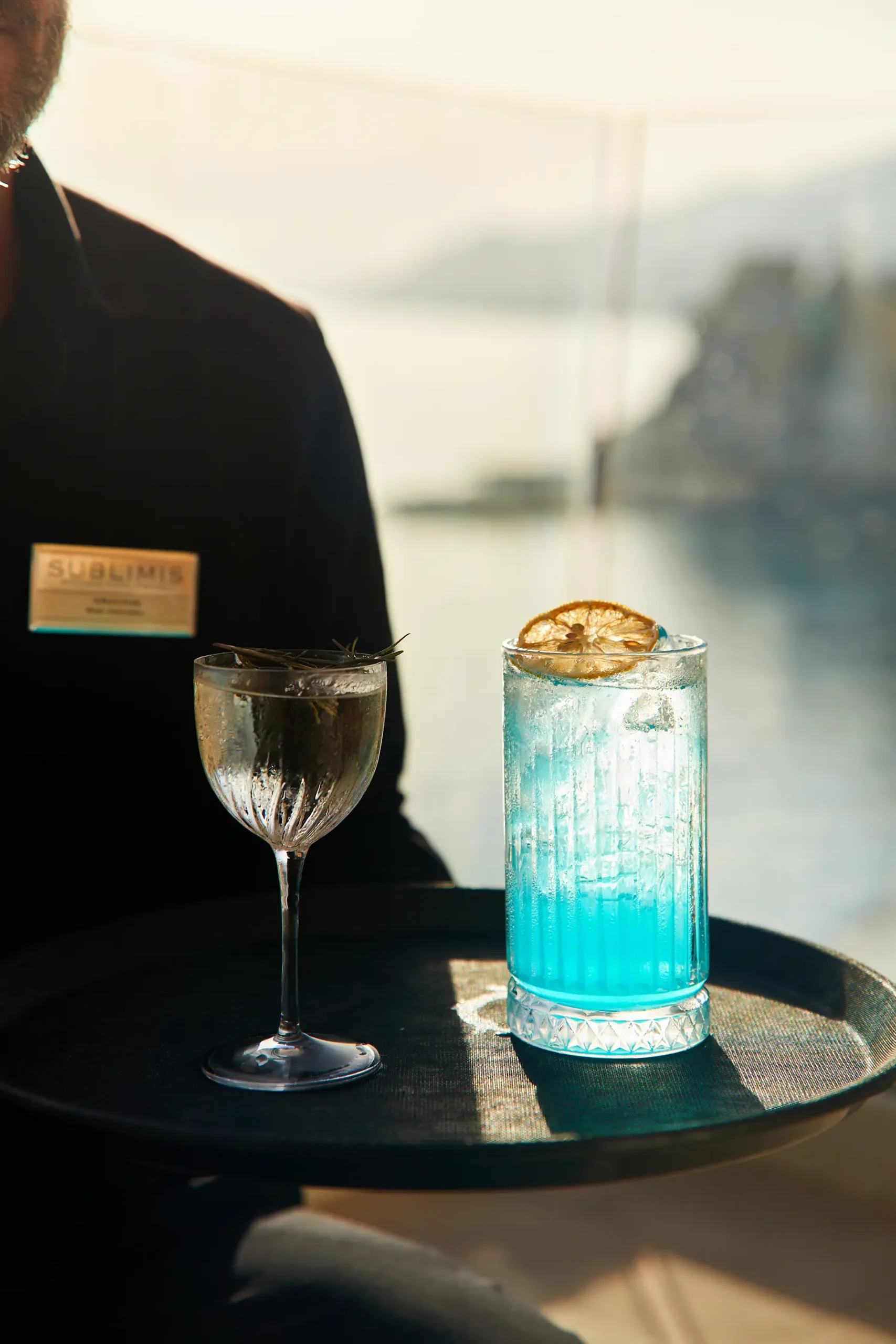 Vassoio nero con due cocktail, uno trasparente in calice e uno azzurro con ghiaccio e fetta di agrume, serviti davanti a una vetrata affacciata sul mare. Sullo sfondo si intravedono la costa e le colline sfocate, illuminate dalla luce calda del sole.
