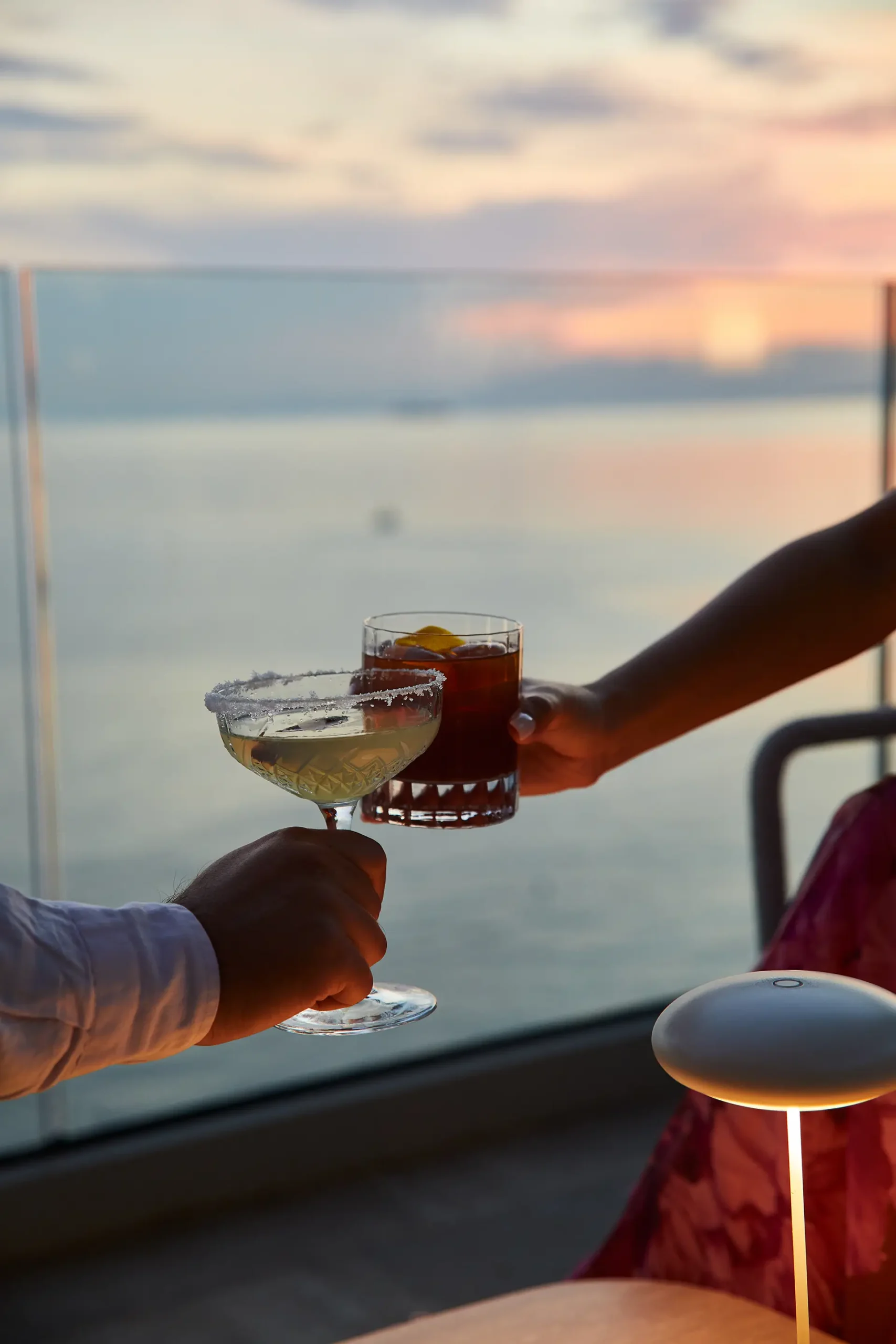Due mani che brindano con cocktail davanti al mare al tramonto, uno chiaro in coppa con bordo zuccherato e uno ambrato con ghiaccio. La luce calda si riflette sull’acqua sullo sfondo, mentre una piccola lampada da tavolo illumina la scena in primo piano.