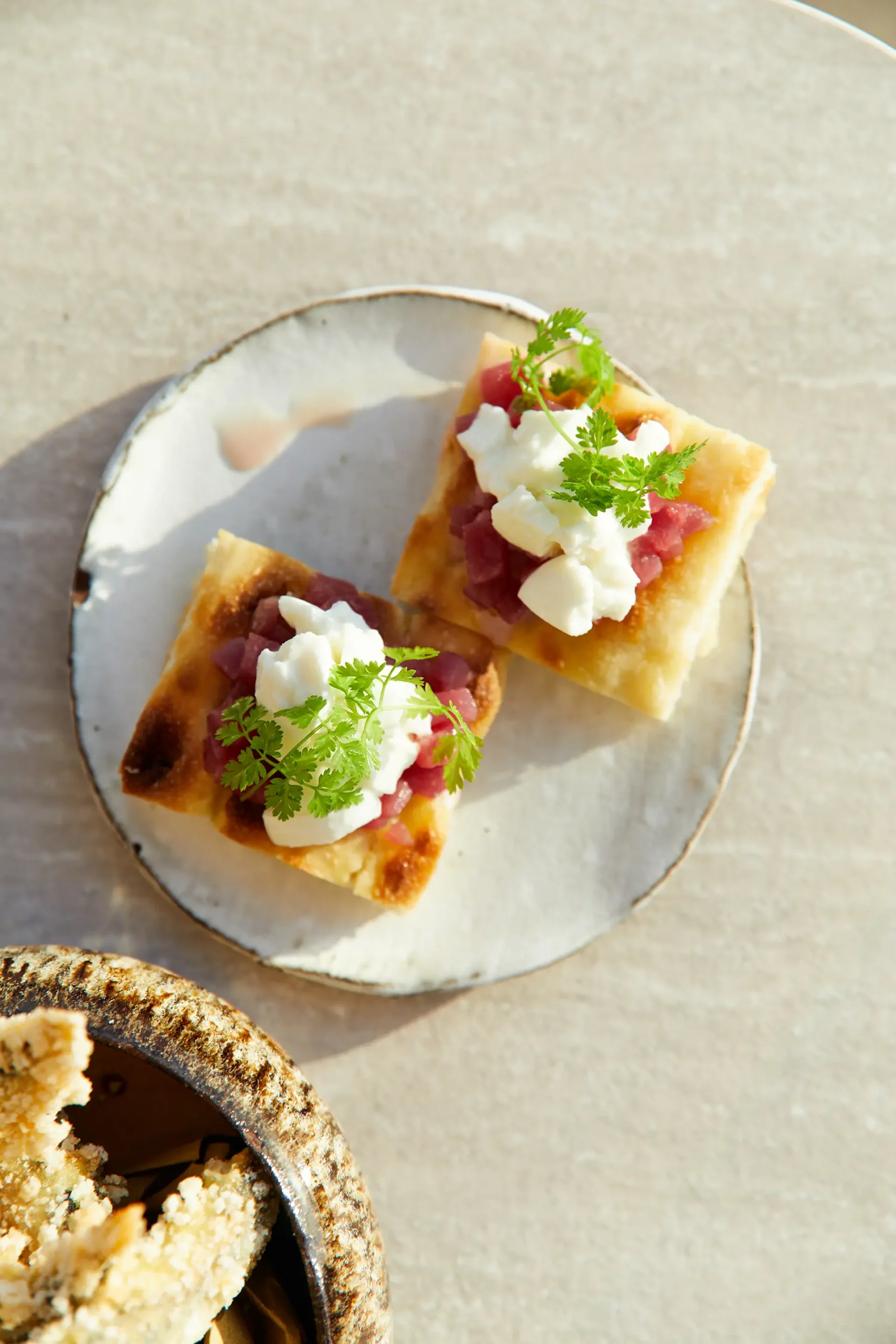 Due tartine servite su un piccolo piatto in ceramica chiara, guarnite con cubetti di tonno, crema bianca e foglie di erbe fresche. La luce calda mette in risalto le texture dorate della base e il contrasto tra i colori degli ingredienti su una superficie neutra.