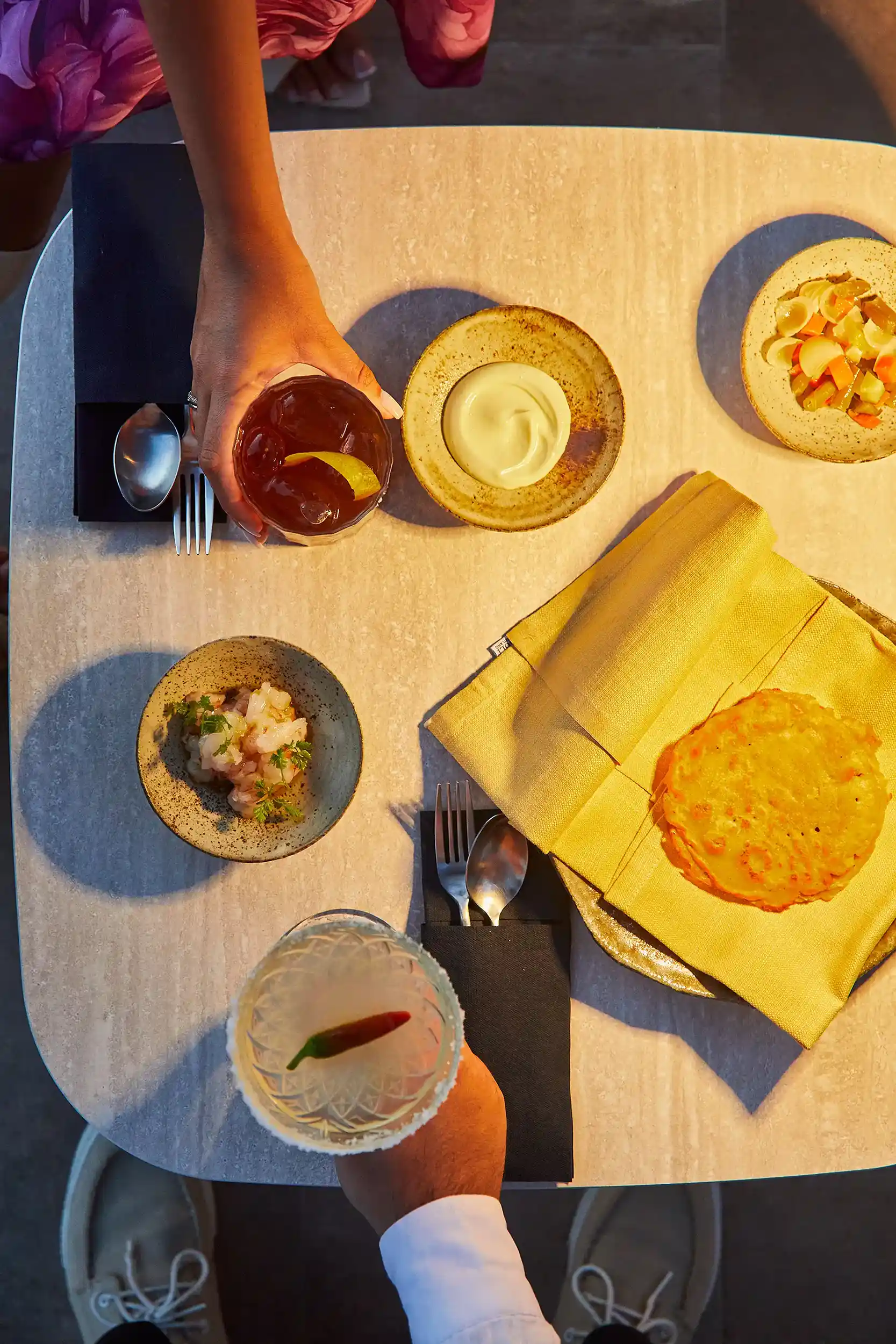 Vista dall’alto di un tavolo apparecchiato con piatti gourmet e dessert: una crema servita in una ciotola chiara, tartine, una preparazione di pesce, pani croccanti e un dessert dorato su tovagliolo giallo. Due mani tengono cocktail, uno ambrato con scorza di agrume e uno chiaro in coppa, mentre le posate e i dettagli della tavola creano una composizione curata e luminosa.