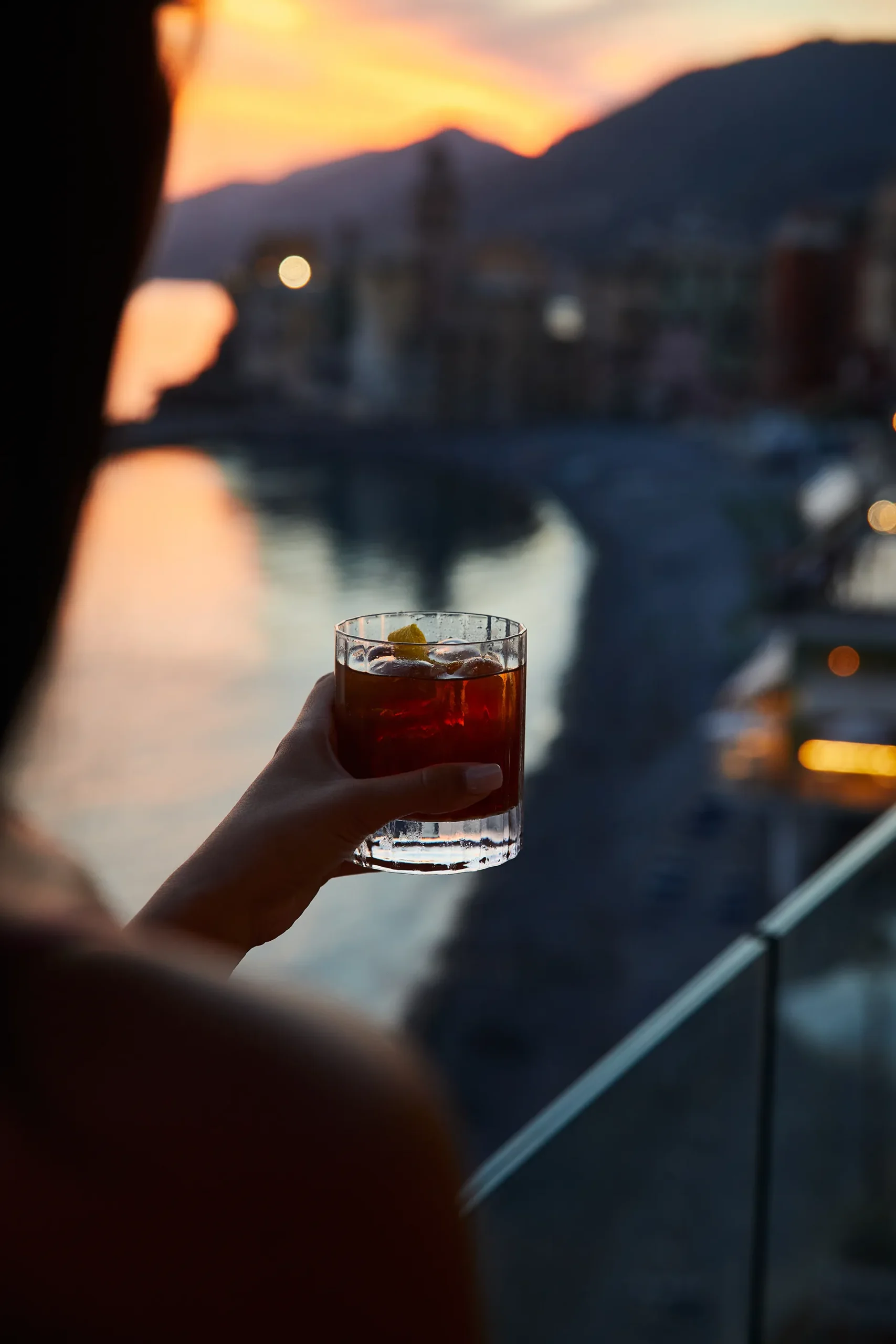 Mano che solleva un bicchiere con cocktail ambrato e ghiaccio davanti a un borgo costiero illuminato dal tramonto. Sullo sfondo sfocato si vedono la spiaggia, Camogli e le montagne, mentre la luce calda si riflette sull’acqua creando un’atmosfera suggestiva.