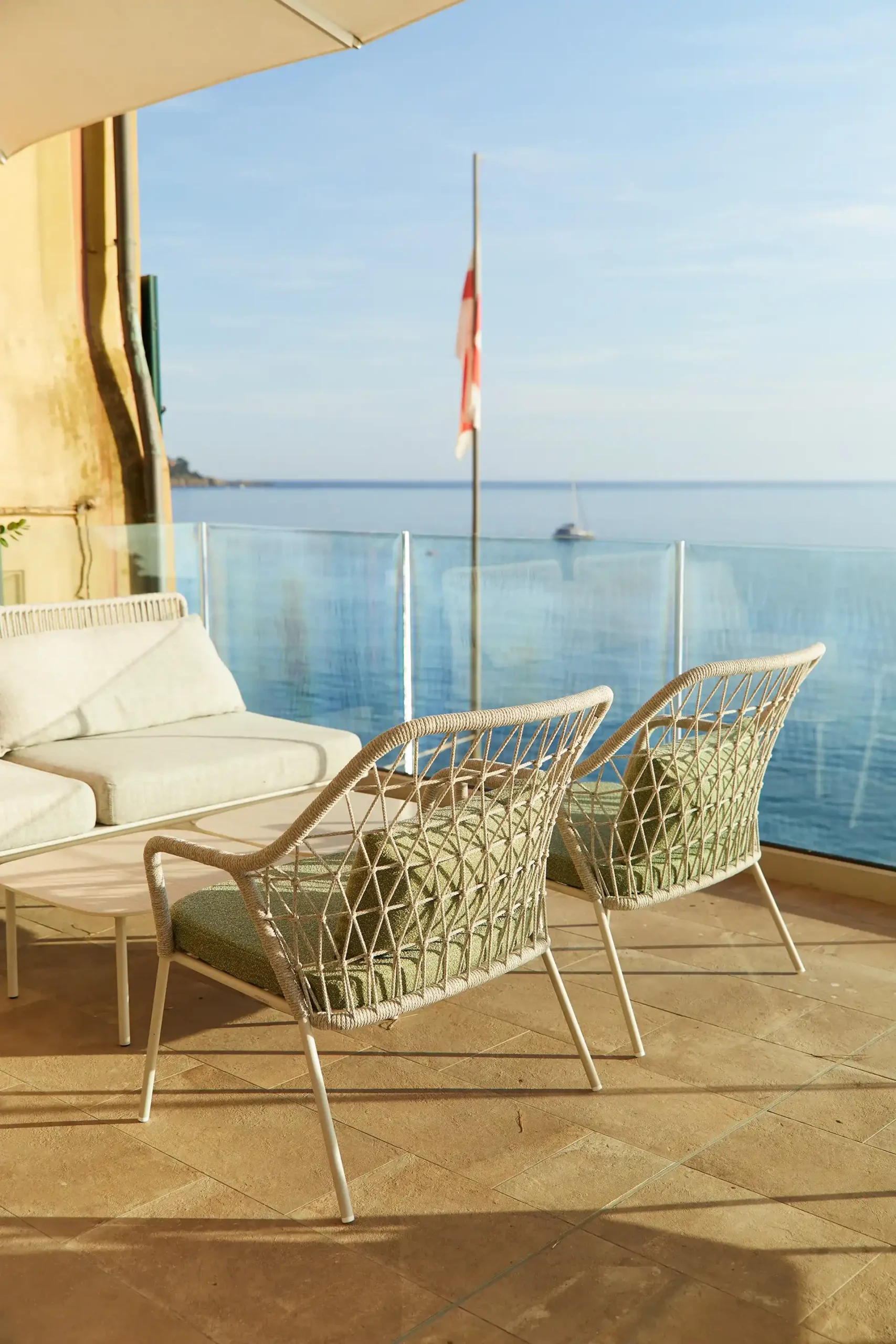 Terrazza affacciata sul mare con due poltroncine intrecciate e un divano chiaro disposti attorno a un tavolino. Una balaustra in vetro offre vista sull’acqua calma, mentre sullo sfondo si vede una bandiera su asta e una barca all’orizzonte sotto una luce calda e dorata.