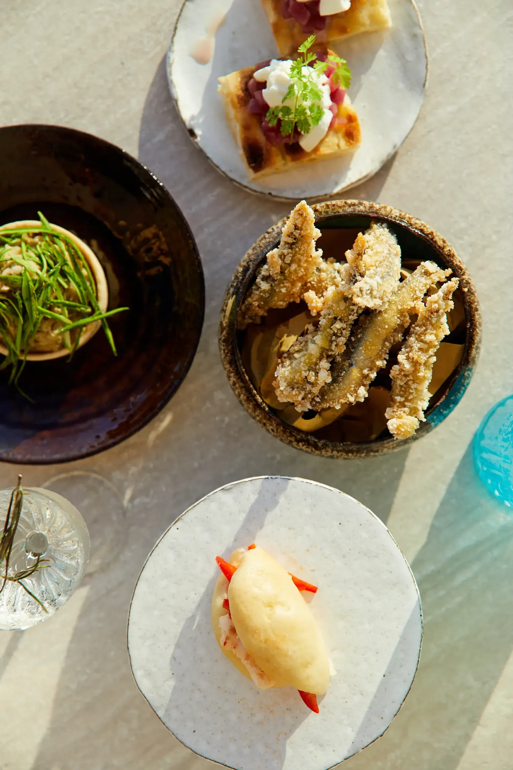 Vista dall’alto di un tavolo apparecchiato con piatti gourmet serviti in ceramiche artigianali: fritti croccanti in una ciotola scura, una quenelle chiara con verdure, una tartina guarnita e una preparazione con crema e fili verdi. Bicchieri e dettagli trasparenti completano la composizione illuminata da luce naturale.